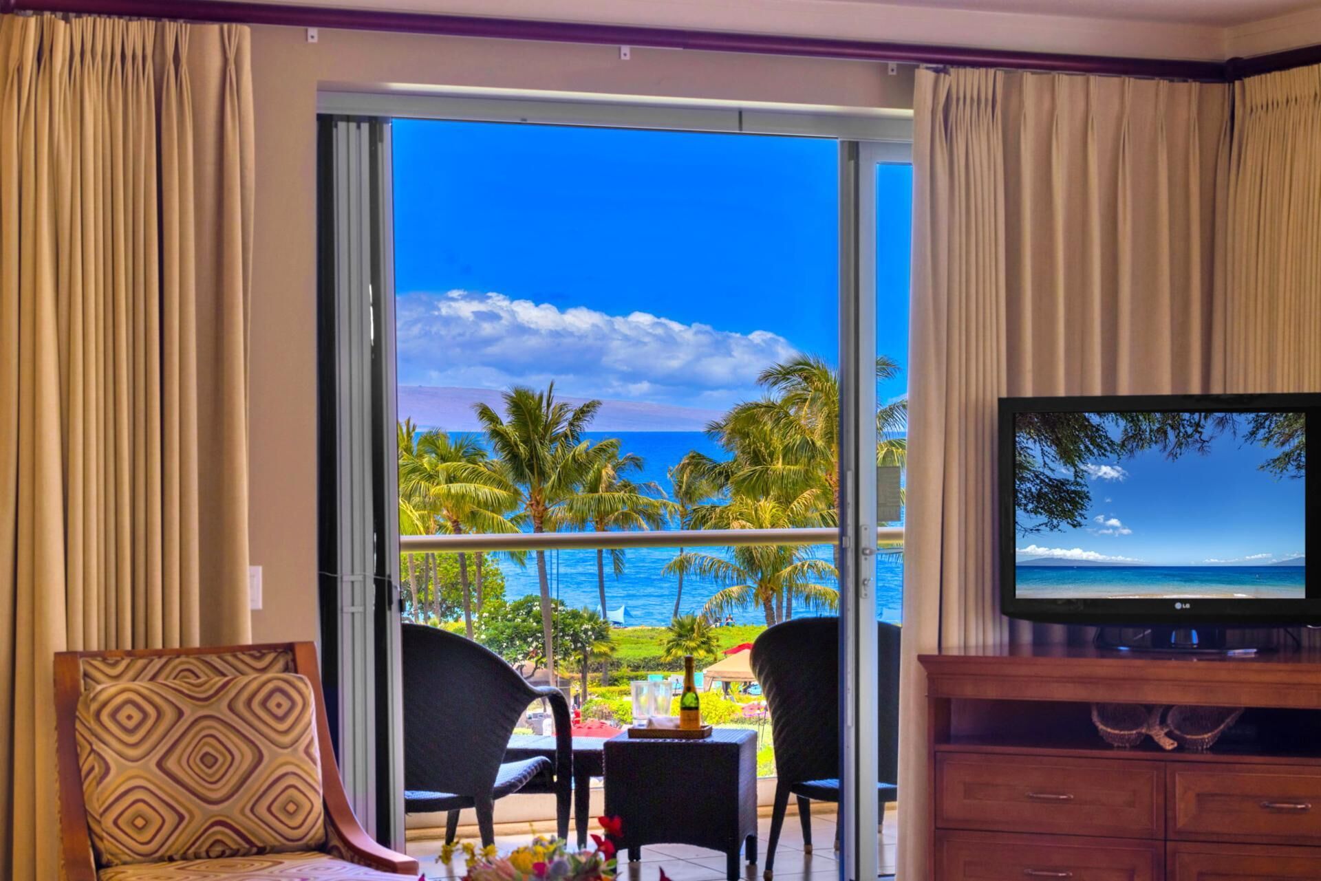 A room with a large sliding glass door leading to a balcony with a view of the ocean and palm trees. There is a television on a wooden cabinet to the right.