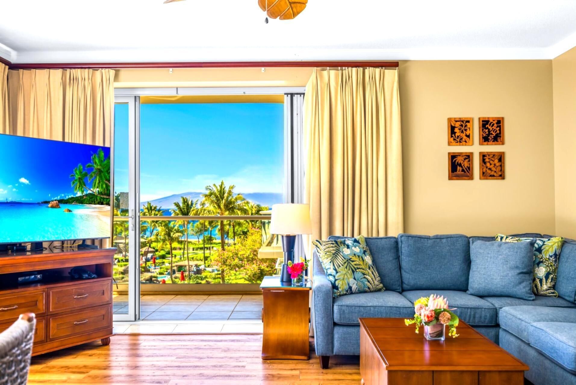 A living room with a large TV, a sofa, a coffee table, and a view of a tropical landscape through a sliding door.