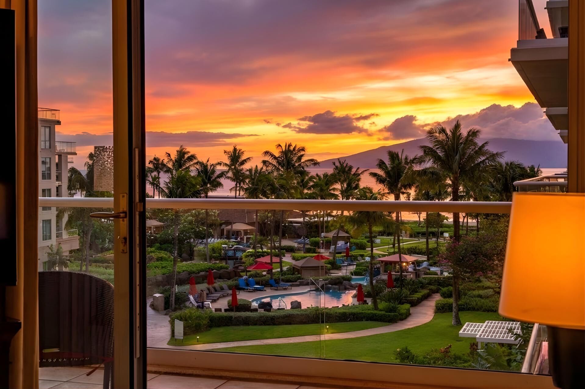 Vista desde una habitación de hotel con balcón que da a una zona turística, con piscina, palmeras y un atardecer.