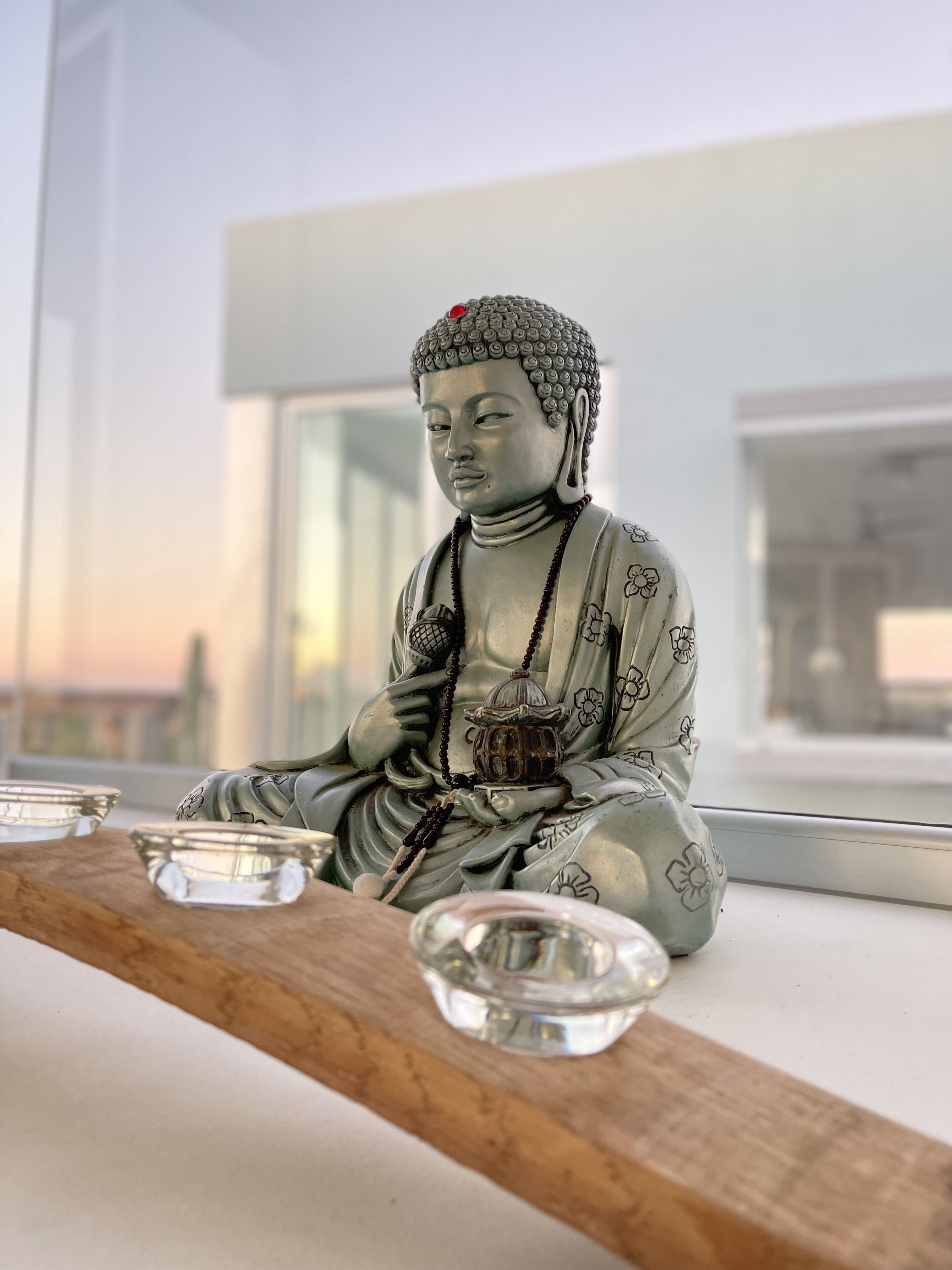 A statue of a seated figure with a beaded necklace and a small lantern, placed on a wooden ledge with three clear glass bowls in front.