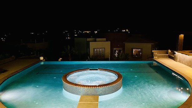 Ein Schwimmbecken mit einem Whirlpool in der Mitte, umgeben von einer gefliesten Terrasse und einem Steinrand.