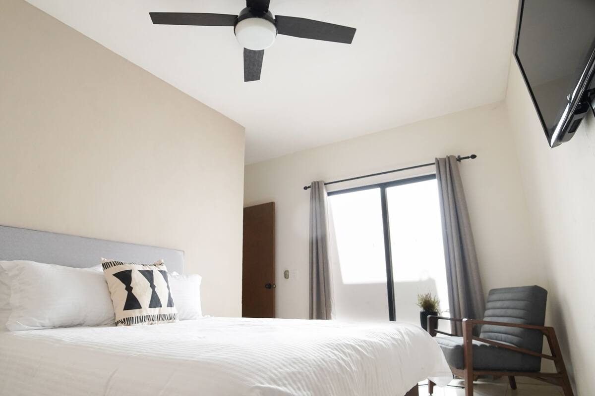 A hotel room with a bed, a ceiling fan, a TV mounted on the wall, a window with curtains, and a chair.