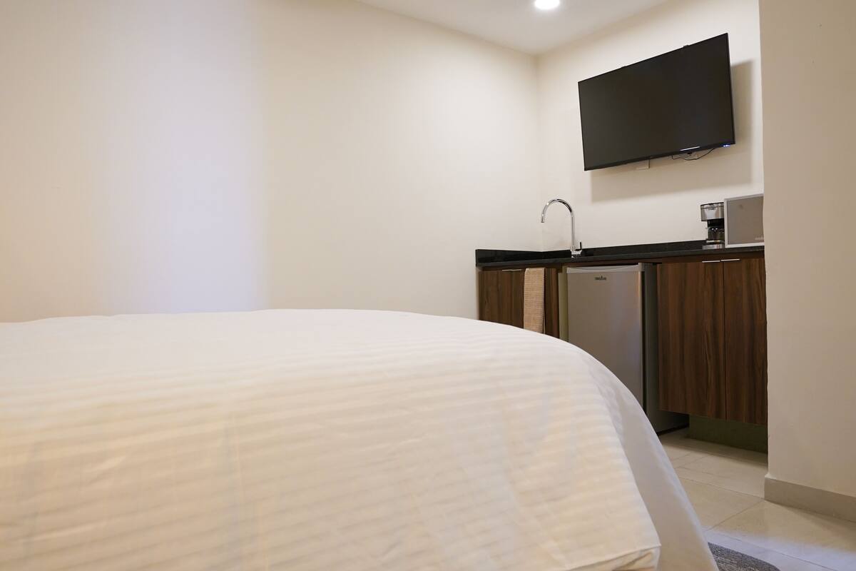 A hotel room with a bed, a flat-screen TV mounted on the wall, a kitchenette with a coffee maker, and a dishwasher.