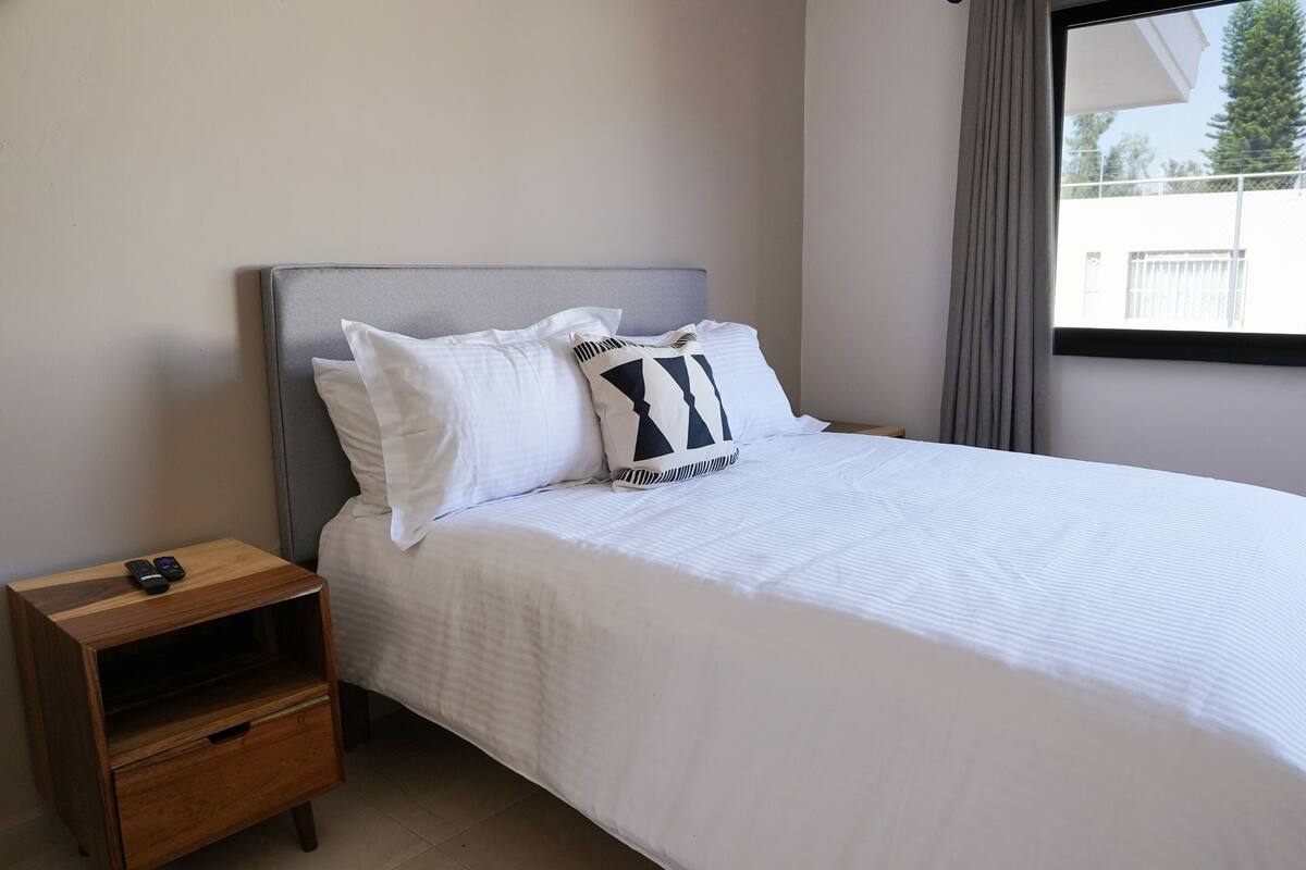 A neatly made bed with white bedding and two pillows, a wooden nightstand with a drawer, and a window with a view of the outside.