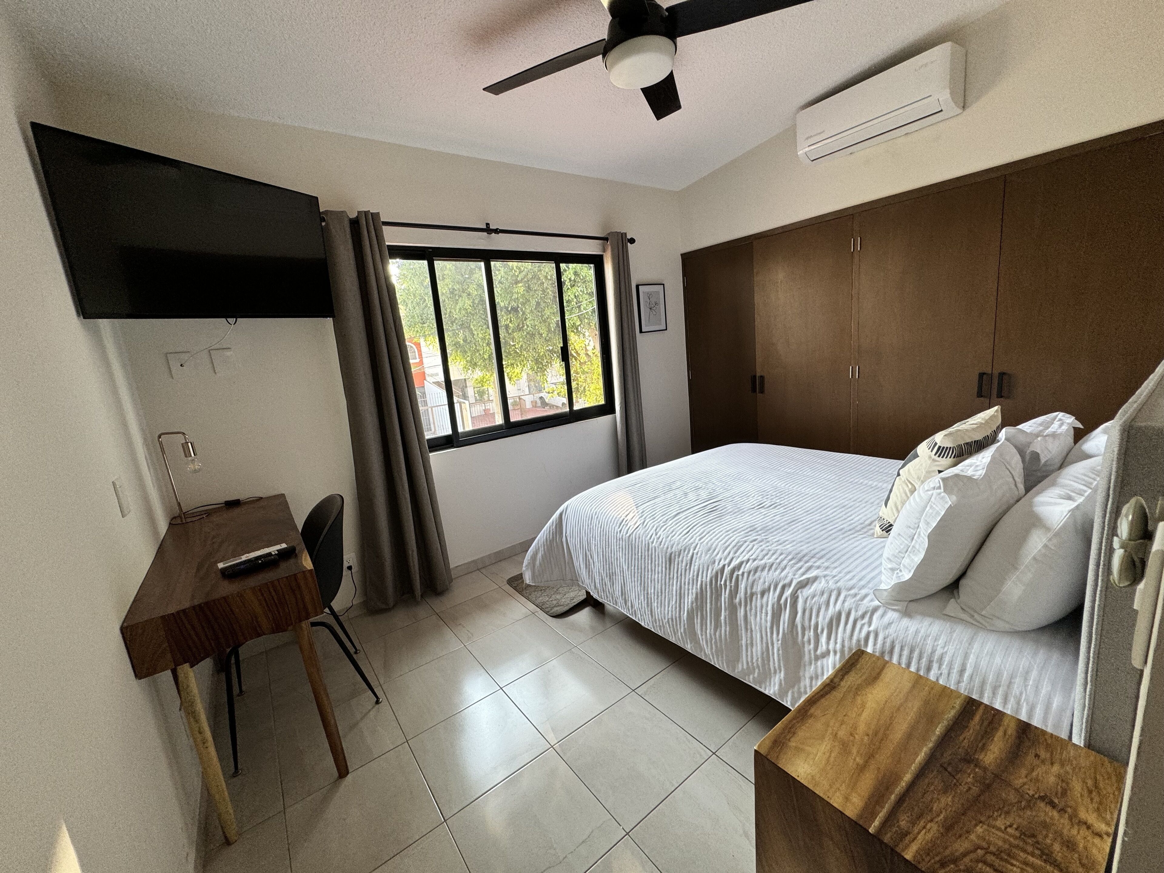 Un dormitorio con cama, escritorio, silla, ventilador de techo y ventana con cortinas.