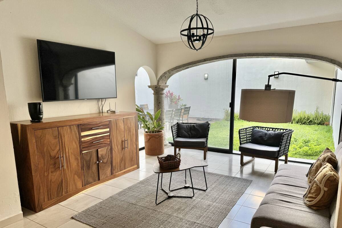 A modern living room with a flat-screen TV, wooden cabinet, and a large window leading to a patio with outdoor furniture.