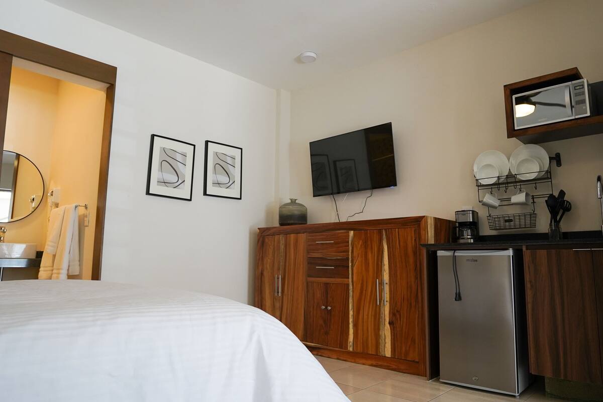 A bedroom with a bed, a wooden wardrobe, a small kitchenette with a microwave, and a TV mounted on the wall.