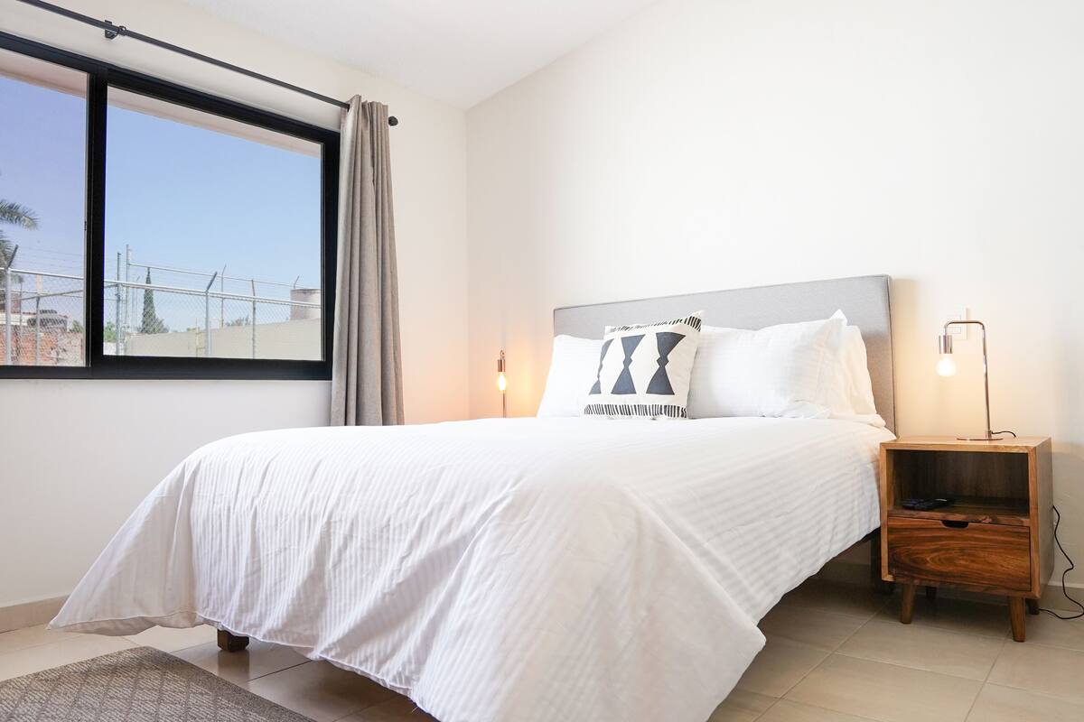A neatly made bed with a white bedspread, a wooden nightstand with a lamp, and a window with a view of buildings and a fence.