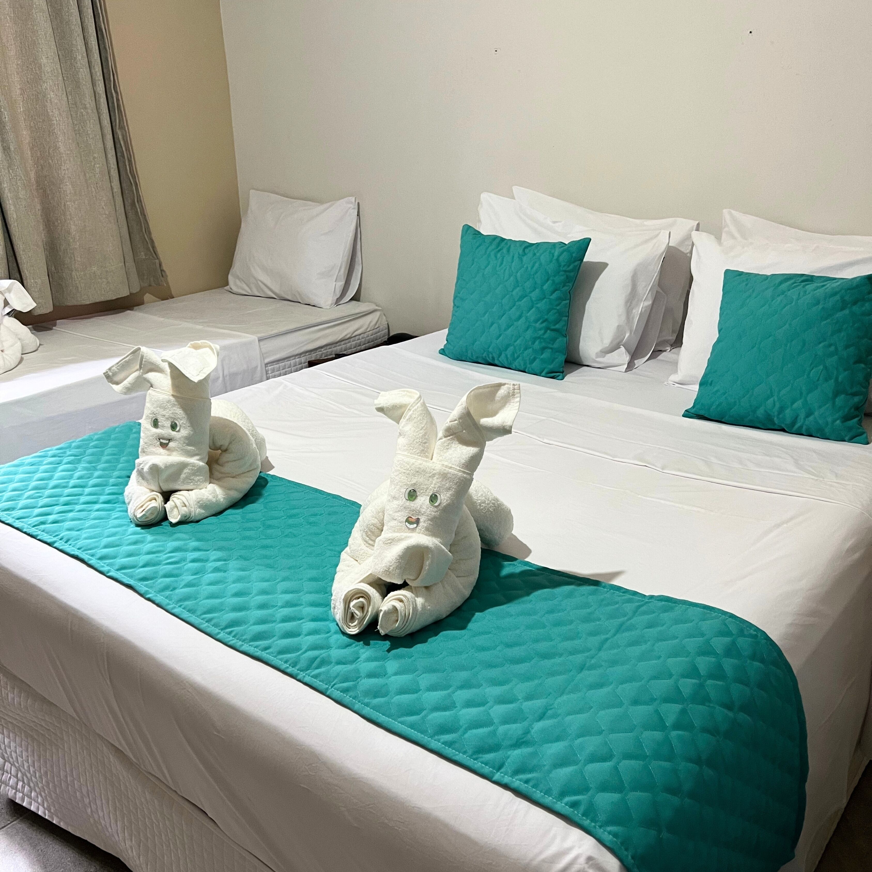 A hotel room with two beds, teal bedding, and two white towels shaped like rabbits.
