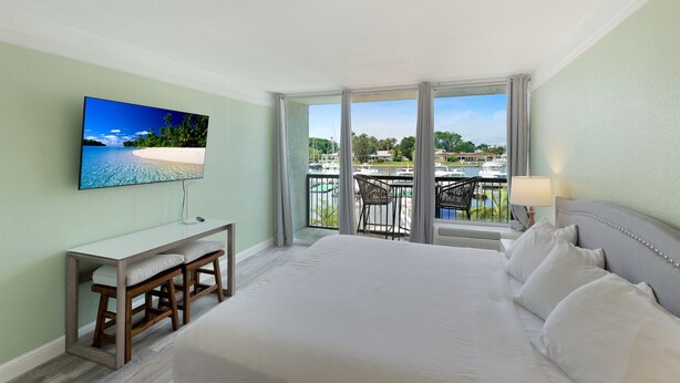 A hotel room with a large bed, a desk with a chair, a TV mounted on the wall, and a balcony with a view of the beach and boats.