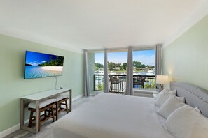 Habitació d'hotel amb un llit gran, escriptori amb cadira, televisió penjada a la paret i un balcó amb vista a la platja i barques.