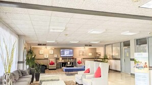 A spacious lobby with a modern design, featuring a large seating area with sofas and armchairs, a flat-screen TV mounted on the wall, and a reception desk.