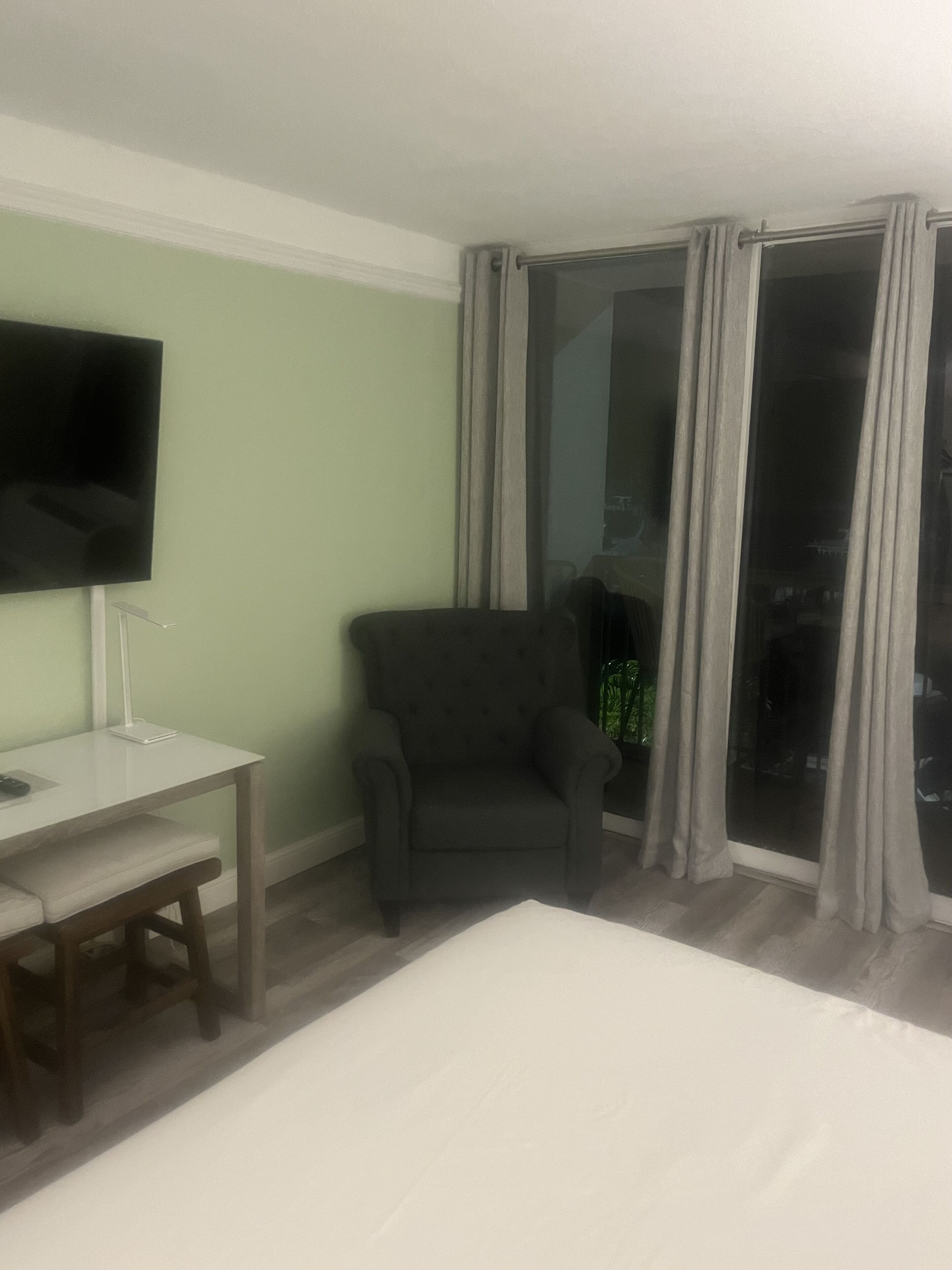 A hotel room with a bed, a desk, a chair, a TV mounted on the wall, and a large window with curtains.