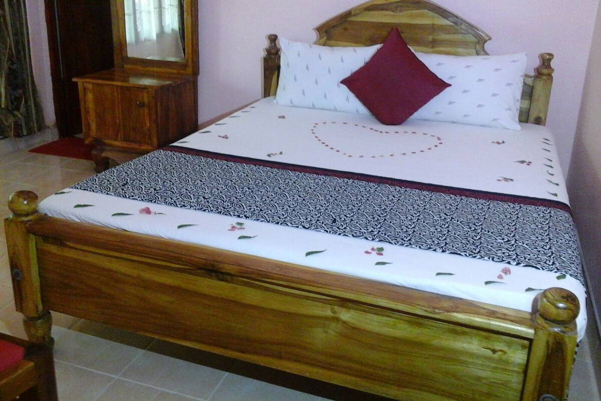 Ein hölzernes Bett mit weißer, gemusterter Bettdecke, einem roten herzförmigen Kopfkissen und einem Spiegel darüber.