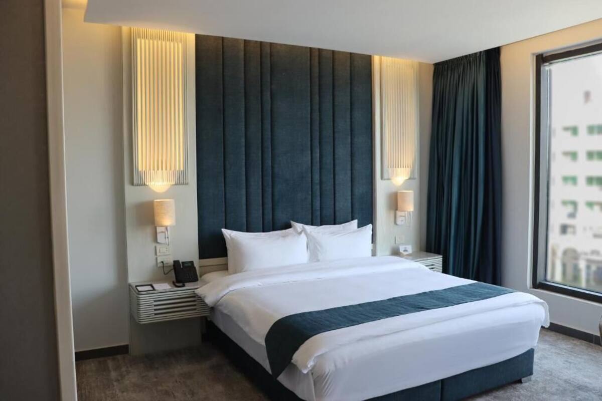 Una moderna camera d'albergo con un letto grande, due comodini con lampade, un telefono e vista sulla città attraverso un'ampia finestra.
