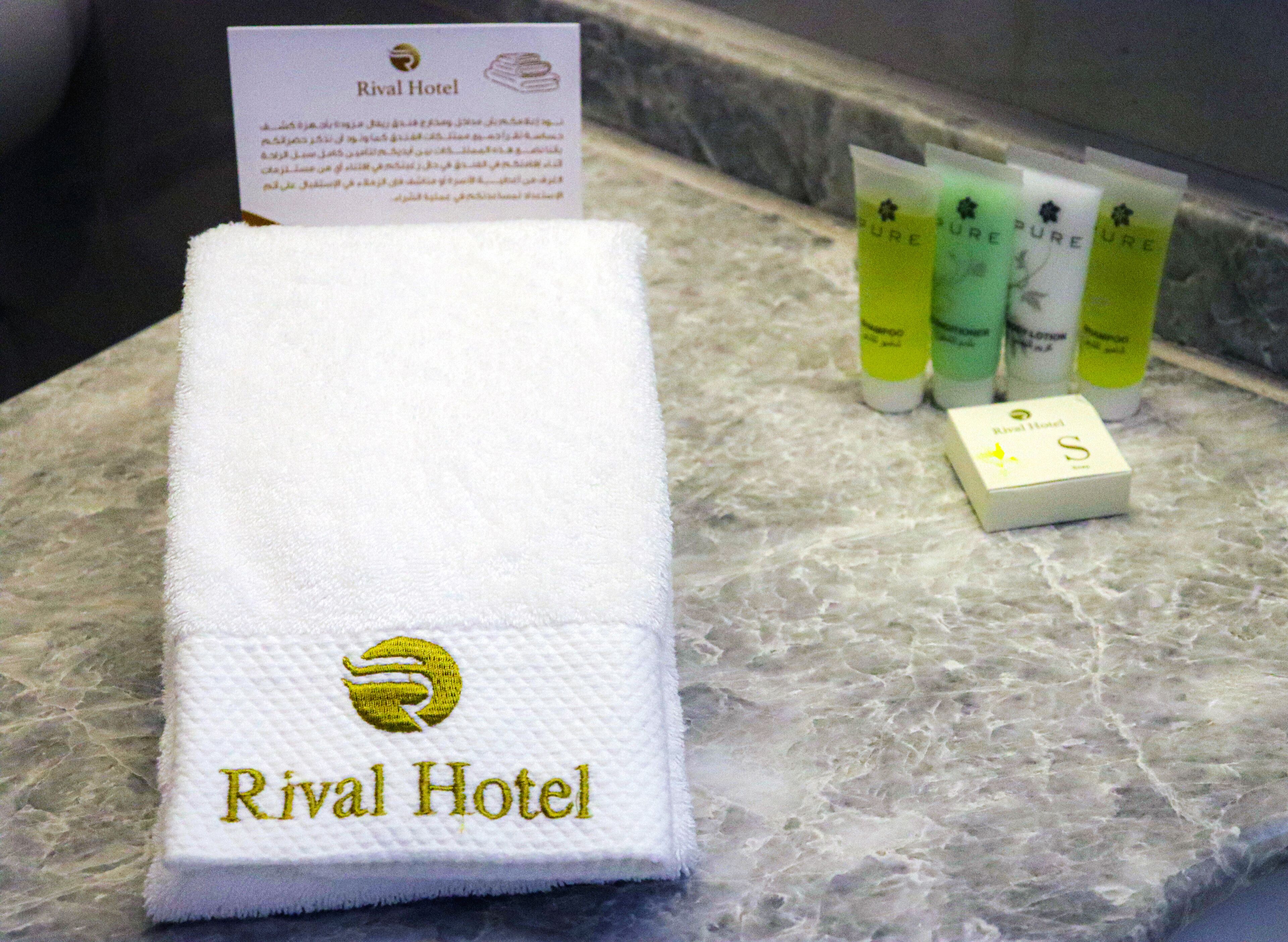 A white towel with embroidered text "Rival Hotel" on a marble surface, accompanied by toiletries.