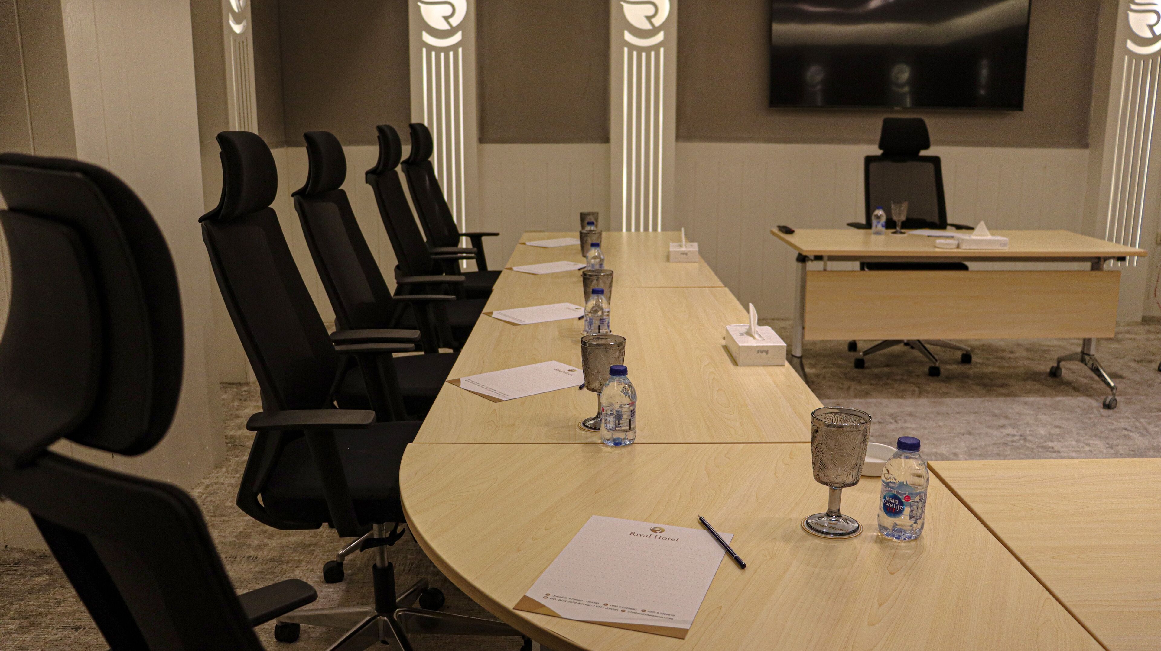Une salle de conférence avec une longue table, des chaises noires, des bouteilles d’eau, une boîte de mouchoirs et un verre posé sur la table.