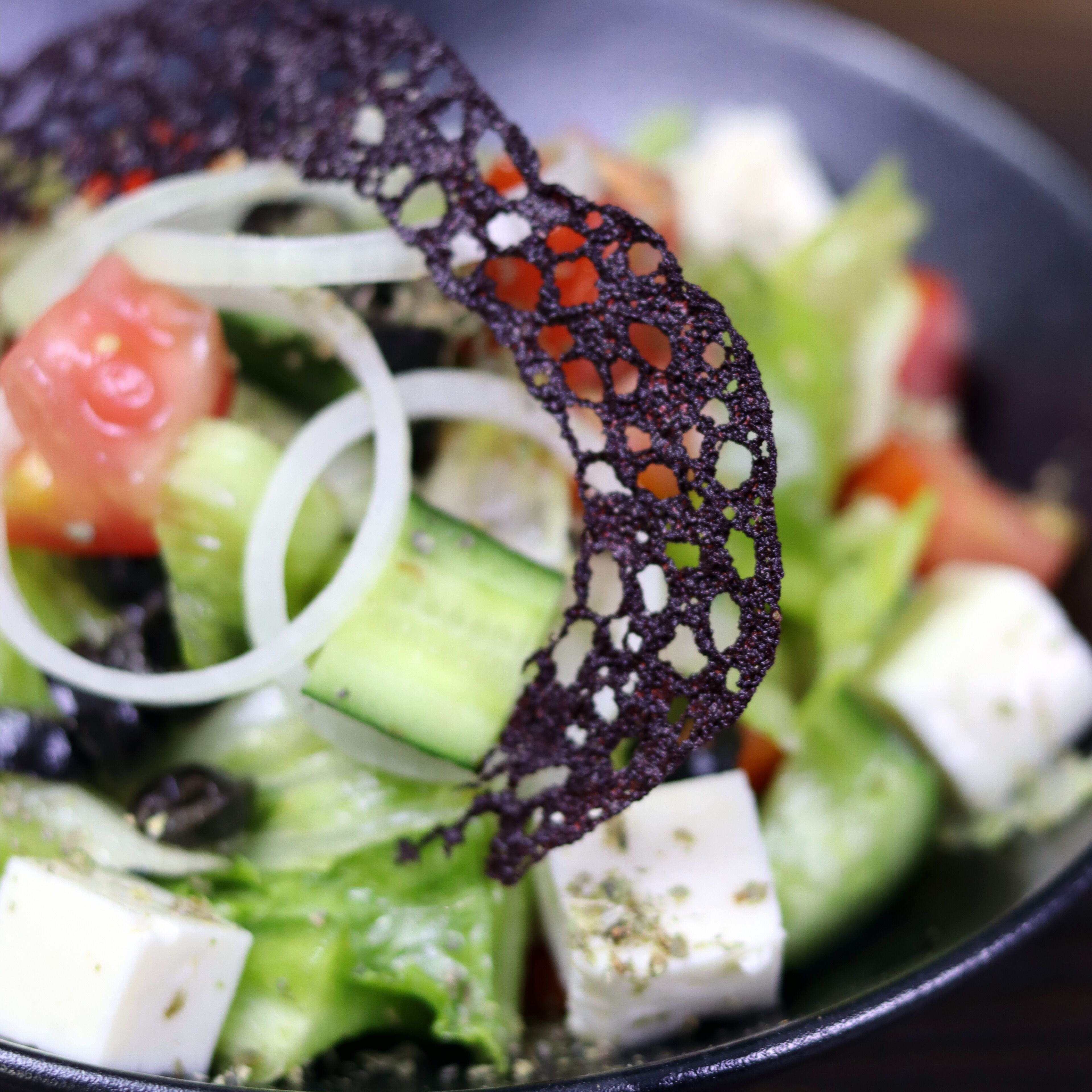 Close-up de uma salada com queijo feta, pepino, tomate e uma renda preta decorativa.
