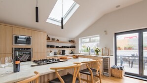 A modern kitchen with wooden cabinets, a built-in oven, a microwave, and a large island with bar stools. - Little Beach House by Bloom Stays -  a holiday let that sleeps 8 guests  in 3 bedrooms (Whitstable)