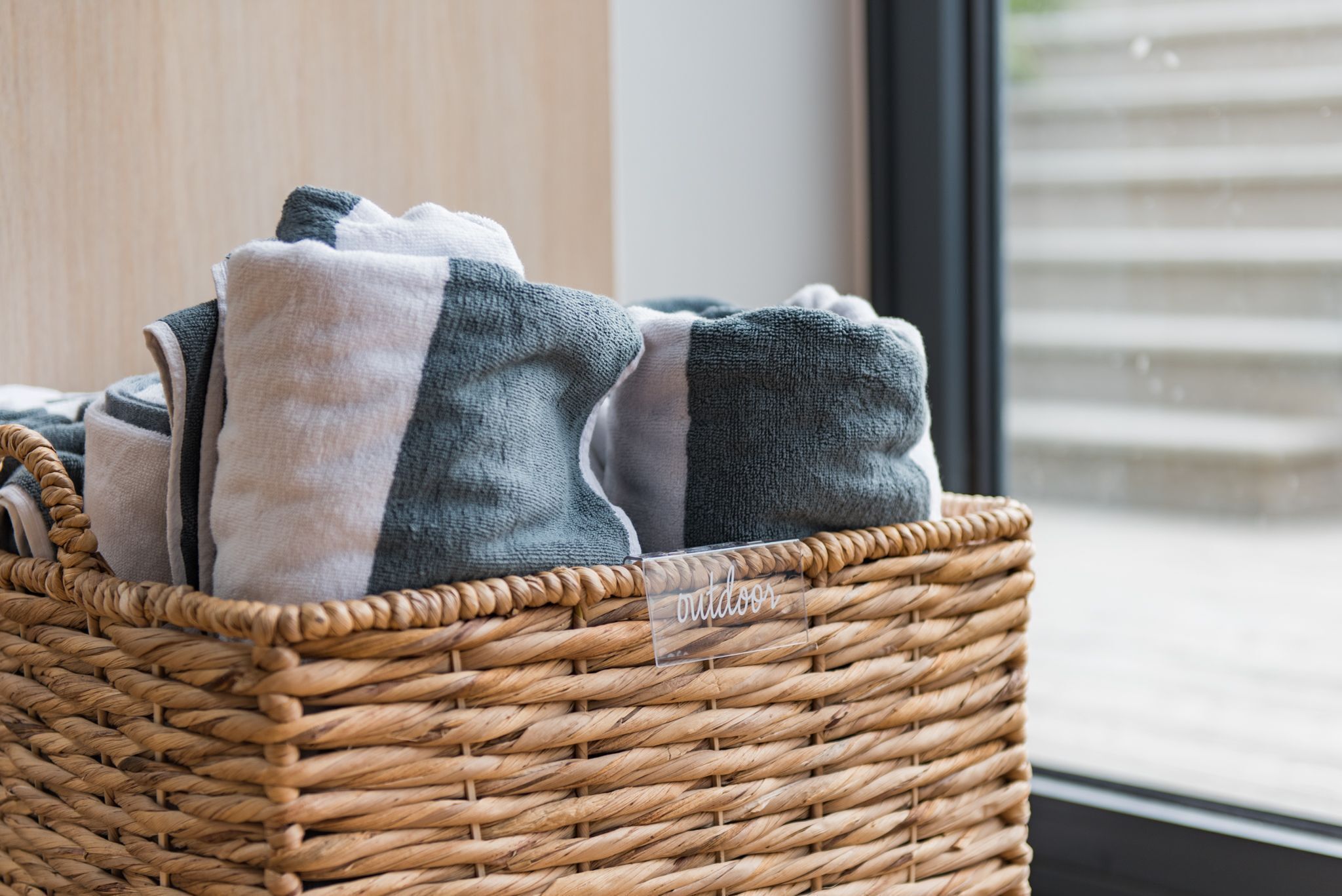 A wicker basket with folded towels labeled "outdoor" and a window in the background.