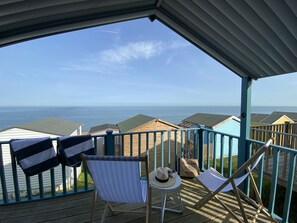 Une terrasse couverte avec vue sur la mer, deux chaises de plage à rayures, un chapeau et une petite table.