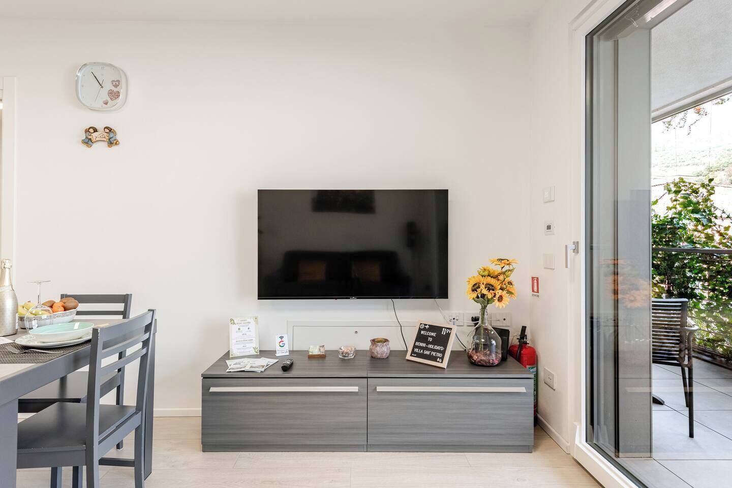Un salon moderne avec un téléviseur à écran plat fixé au mur, un coin repas avec une table et des chaises, et une porte vitrée donnant sur un espace extérieur.