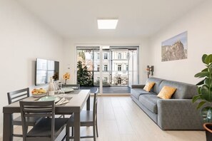 A modern living room with a dining area, a grey sofa, and a large window offering a view of buildings and trees. - BENINI HOLIDAYS: VILLA SAN PIETRO (Arco)