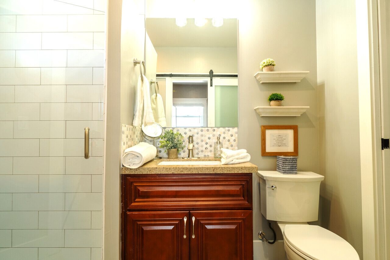 Une salle de bain avec des murs recouverts de carreaux blancs, un meuble-lavabo en bois, un lavabo avec un miroir et des toilettes. On y trouve des serviettes et une petite plante sur le meuble-lavabo.