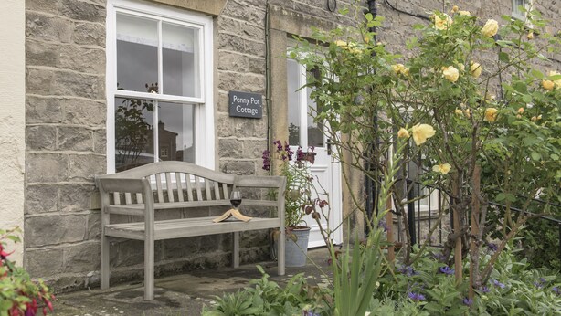 Une petite maison en pierre avec un banc à l’extérieur, une pancarte indiquant « Penny Pot Cottage », et un jardin agrémenté de fleurs jaunes et violettes.