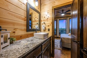 Baño de cabaña de madera con encimera de granito, lavamanos blanco, espejo grande y vista al exterior a través de una puerta.