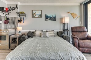 Un dormitorio con cama, un sillón de cuero marrón, una zona de cocina con estufa, y una pared decorada con un mapa y un cuadro.