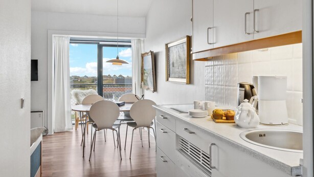 A modern kitchen with white cabinets, a large window, and a dining area with chairs. - 1 bedroom beach front apartment in Fanø (Fanø)