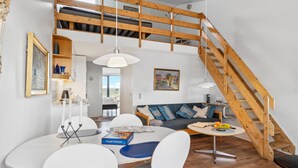 A modern loft-style living space with a wooden staircase, a dining area with a white table and chairs, a sofa with patterned cushions, and a painting on the wall. - 1 bedroom beach front apartment in Fanø (Fanø)