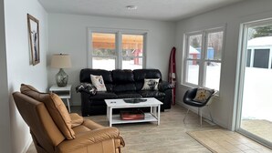 Ein Wohnzimmer mit einem schwarzen Ledersofa, einem braunen Ledersessel, einem weißen Couchtisch und einem roten Surfbrett, das an der Wand lehnt.