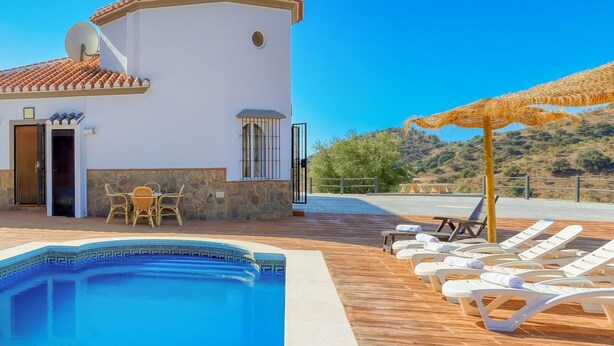 Un área de piscina con sillones de descanso y una mesa, un edificio con fachada blanca y detalles de piedra, y una vista despejada del paisaje circundante.