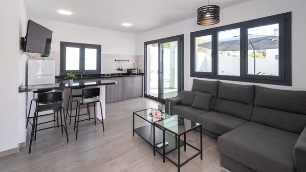 A modern living room with a grey sofa, a glass coffee table, and a kitchen area with a dining set. - Villa La Fuente Periana by Ruralidays (Málaga)
