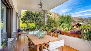 A covered patio with a wooden table and chairs, surrounded by potted plants and a view of a mountainous landscape.