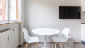 Une cuisine moderne avec une table et des chaises blanches, un téléviseur noir fixé au mur et une fenêtre avec des stores.