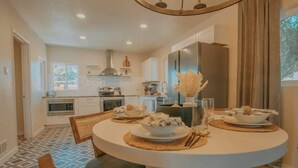 Cozinha moderna com armários brancos, uma mesa de jantar redonda com jogos americanos e vista para o exterior pela janela.