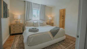 Quarto com uma cama grande, duas mesas de cabeceira com luminárias, uma janela com cortinas e uma porta que dá para outro cômodo.
