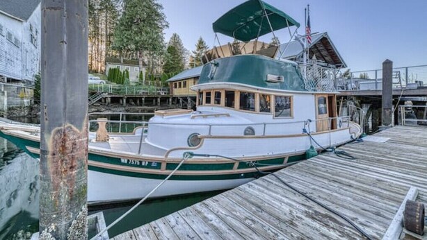 A white boat docked at a wooden pier with a green canopy, surrounded by trees and buildings. - Historic Charming Boat in Downtown Harbor

 (Gig Harbor)