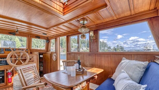 A wooden interior with a dining table, chairs, a sofa, and a view through large windows. - Spacious 46' Yacht: Luxury, kayaks, walk to town

 (Gig Harbor)