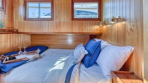 A wooden-paneled room with a bed, two windows, and a nightstand with a tray. - Spacious 46' Yacht: Luxury, kayaks, walk to town

 (Gig Harbor)