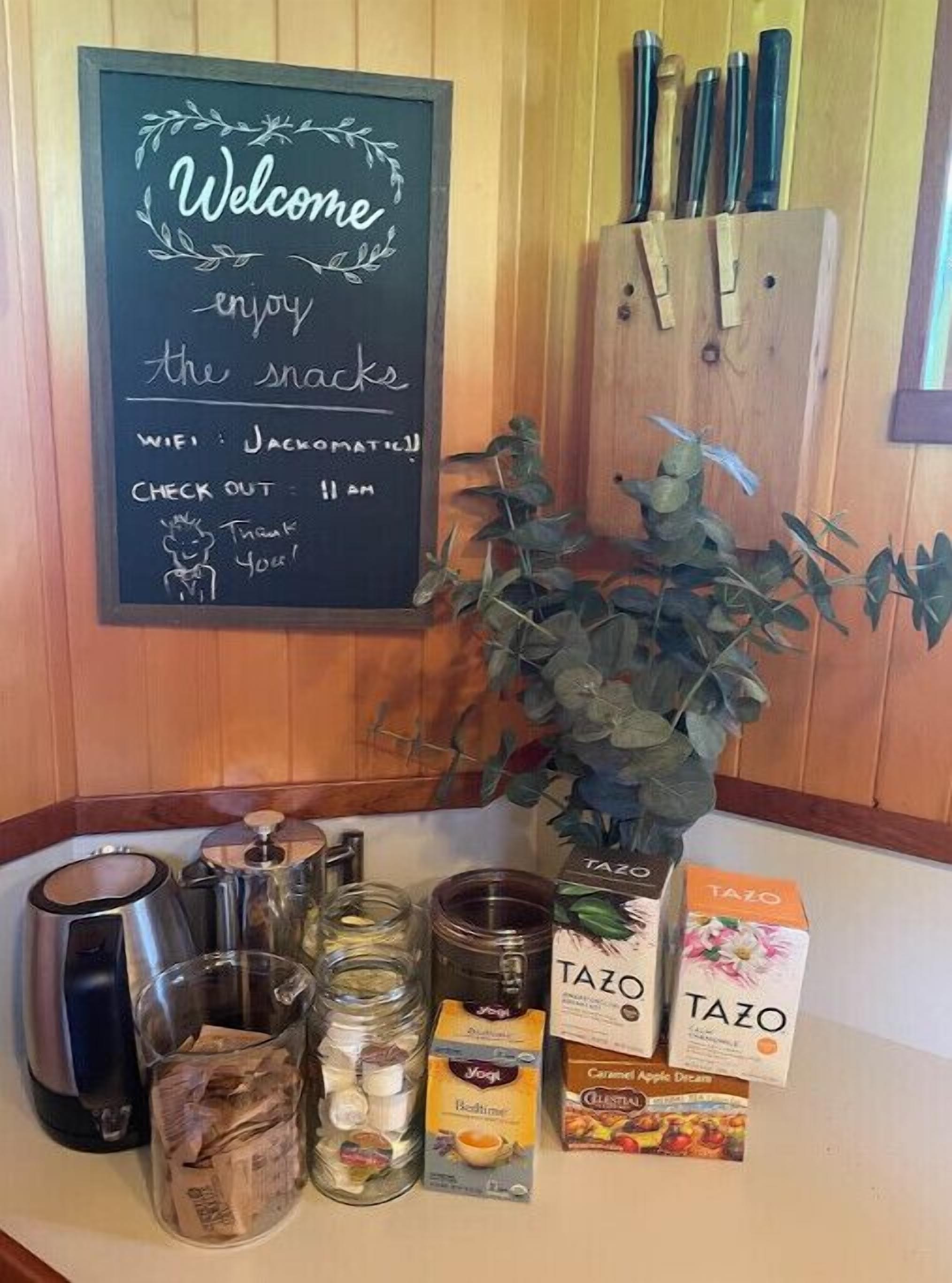 En välkomnande skylt med ett meddelande, en kaffebryggare, en burk med te, en ask med Tazo-te och en växt.
