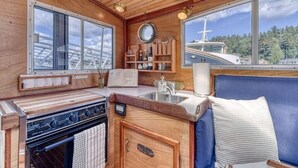 Cocina pequeña con estufa, lavamanos y armarios de madera. Se ve una ventana con vistas a un barco y árboles.