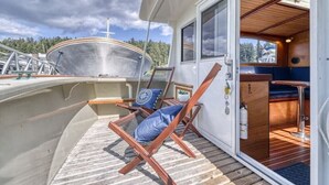 Cubierta de barco con una silla de salón, una mesita y vista al agua y al bosque.