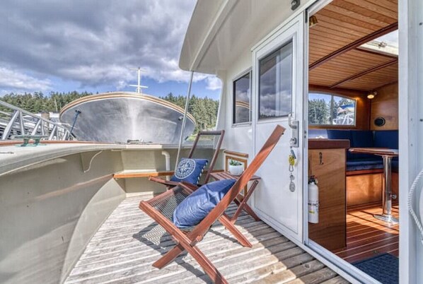 A boat deck with a lounge chair, a small table, and a view of the water and forest.