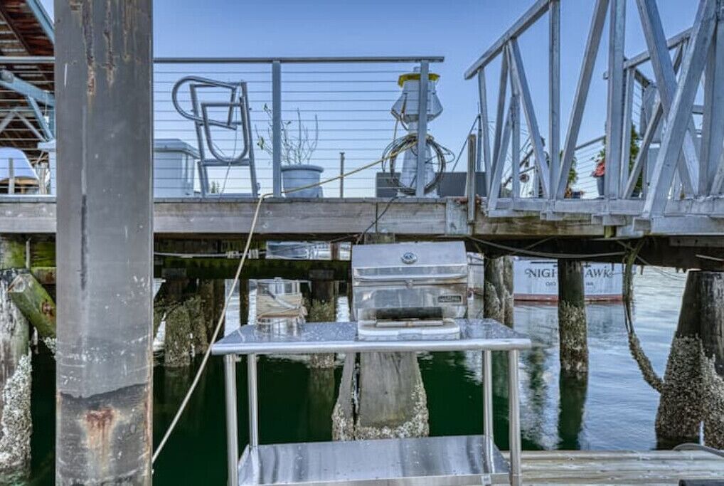 A metal dock structure with a grill and a chair, surrounded by water and wooden posts.