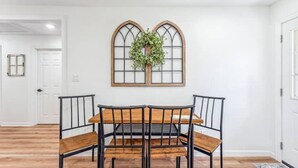 Een eetruimte met een houten tafel en zwarte metalen stoelen, een raam met een krans, en een witte deur op de achtergrond.