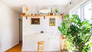 Une cuisine lumineuse avec des murs blancs, un plancher en bois, une table à manger blanche, deux chaises blanches, un luminaire suspendu et une variété de plantes en pot.